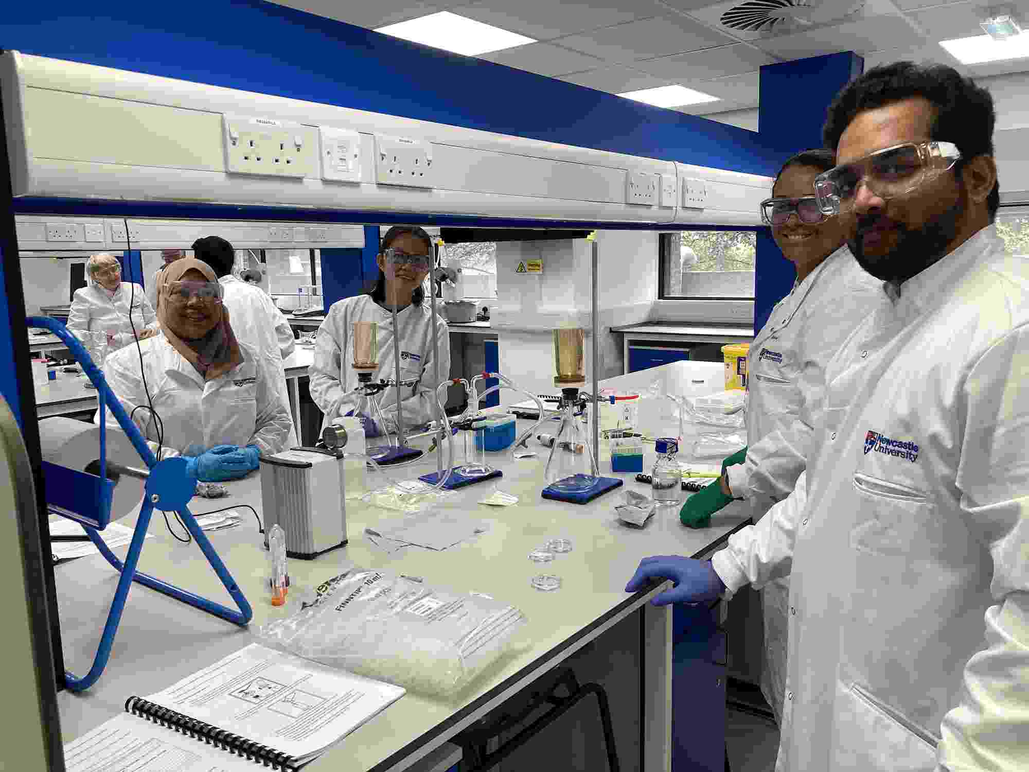 Several people wearing lab coats and safety equipment stand around a work bench in a laboratory, with water sampling equipment spread out on the bench